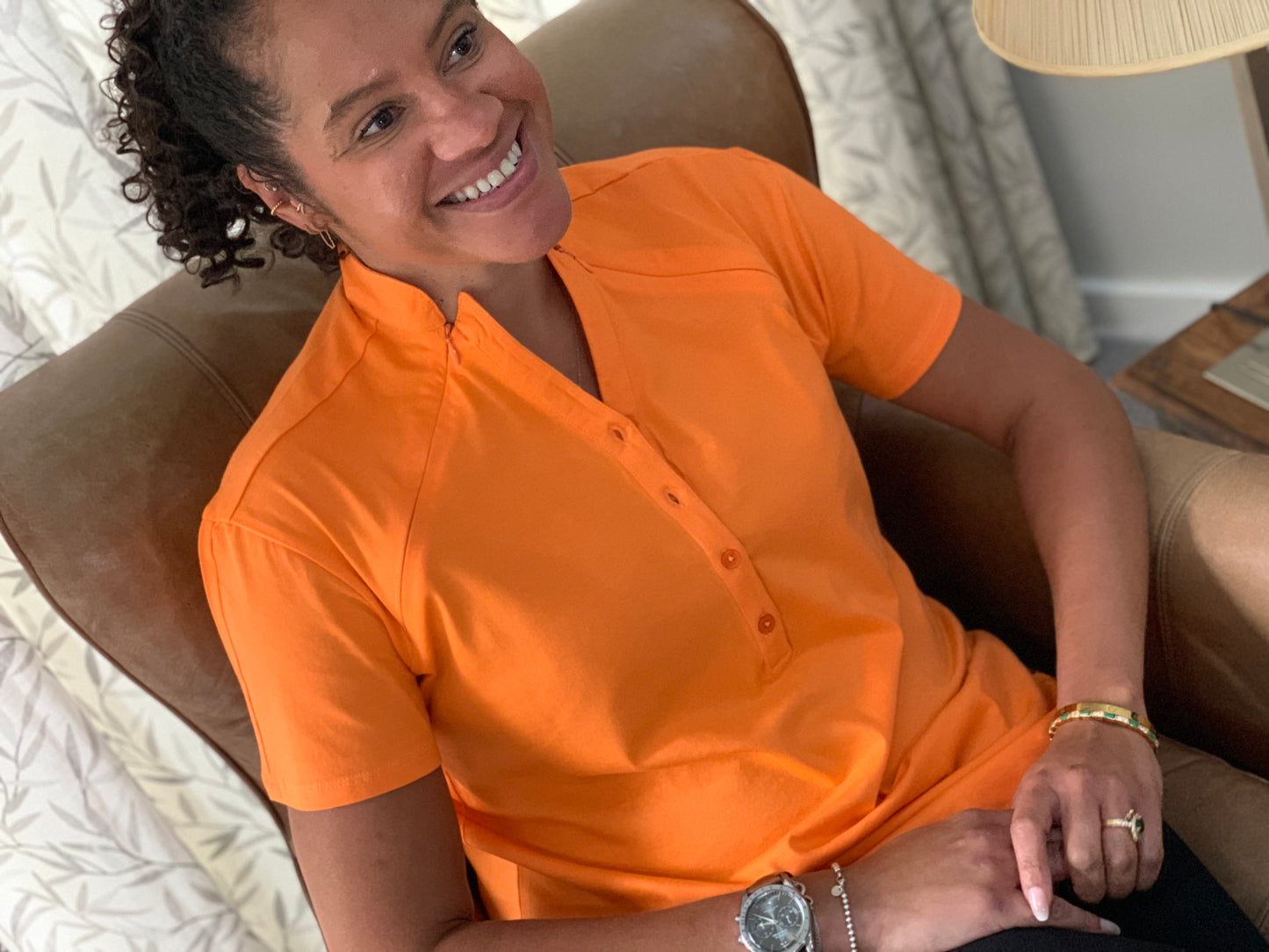 The tangelo orange ChicComfort Chemowear with short sleeves. The model is sitting and smiling in an armchair. The buttons and zips are done up, showing the shape of the collar